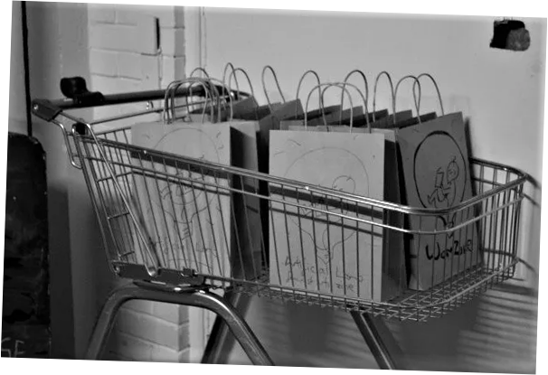 A black and white photograph of a  supermarket shopping trolley filled with paper bags with prints on them that read "Artificial Womb" 