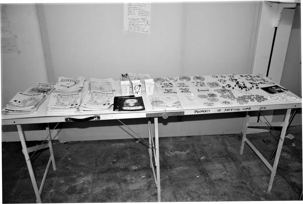 Black and white photograph of a foldable table covered in zines, bracelets and other small trinkets for sale.