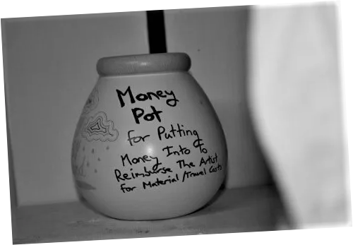 A close up black and white photograph of a small pot. The text on the pot reads "Money Pot. For putting money into to reimburse the artist for material/travel costs"