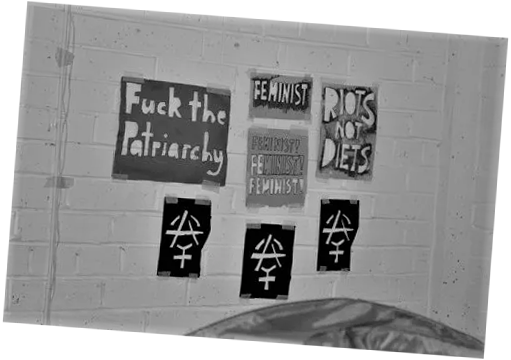 A black and white photograph of a collection of spray paint stencils taped to a brick wall. The stencils read "Fuck the Patriarchy", "FEMINIST", "FEMINIST! FEMINIST! FEMINIST!", and "RIOTS NOT DIETS". There are also 3 stencils with the anarcha-feminist symbol on them.