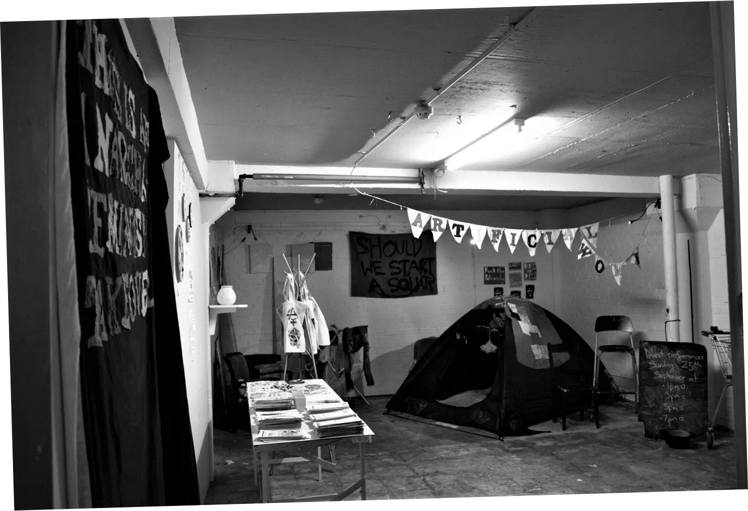 Black and white photograph of exhibition set up for 'This is an Anarcha-Feminist Takeover'. A tent features prominently alongside a clothes horse with clothes hanging from it, bunting with the words 'ARTIFICIAL WOMB' sewn onto it, and spray paint stencils and a large piece of fabric with 'SHOULD WE START A SQUAT?' spray painted on it are hung on a wall in the background. A blackboard sign in the foreground reads 'Performance: 6.30pm, 7.30pm. BYOB' in chalk. To the right there is the shop table, printed tote bags on a clothes hanger, and the money pot on a shelf.