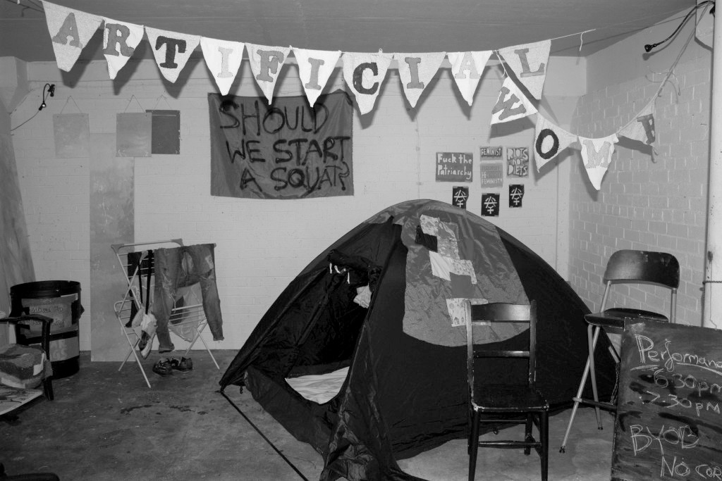 Black and white photograph of exhibition set up for 'This is an Anarcha-Feminist Takeover'. A tent features prominently alongside a clothes horse with clothes hanging from it, bunting with the words 'ARTIFICIAL WOMB' sewn onto it, and spray paint stencils and a large piece of fabric with 'SHOULD WE START A SQUAT?' spray painted on it are hung on a wall in the background. A blackboard sign in the foreground reads 'Performance: 6.30pm, 7.30pm. BYOB' in chalk.