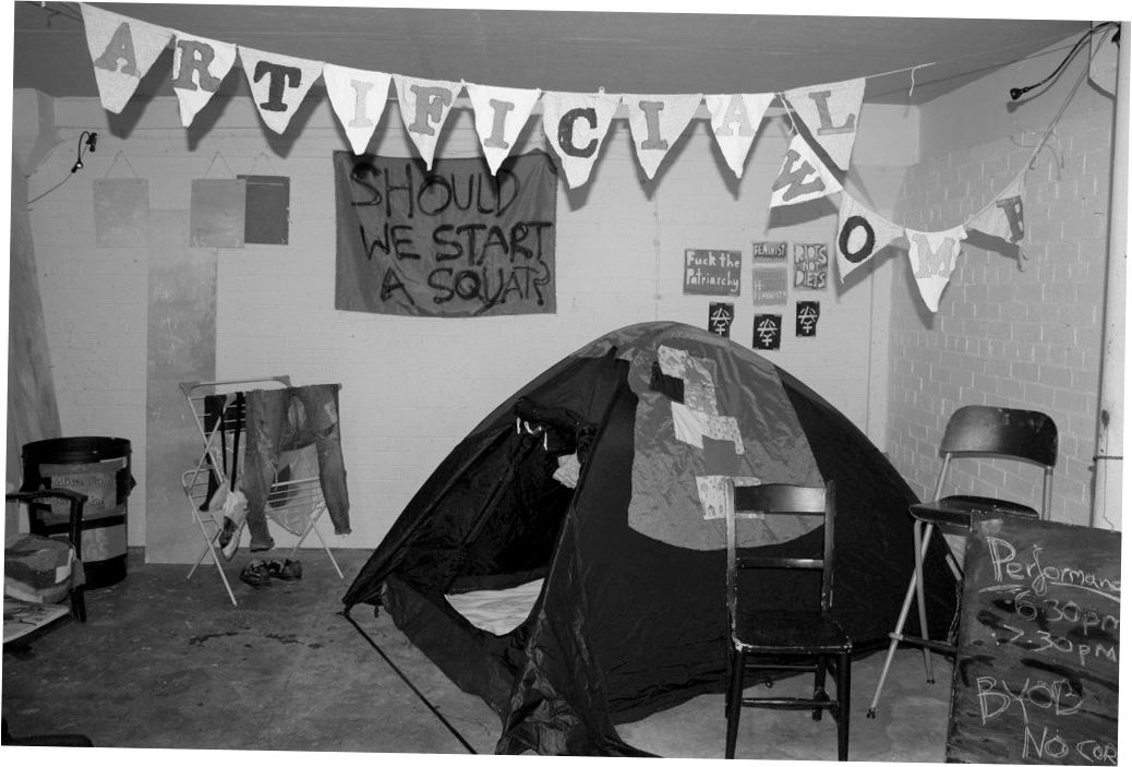 Black and white photograph of exhibition set up for 'This is an Anarcha-Feminist Takeover'. A tent features prominently alongside a clothes horse with clothes hanging from it, bunting with the words 'ARTIFICIAL WOMB' sewn onto it, and spray paint stencils and a large piece of fabric with 'SHOULD WE START A SQUAT?' spray painted on it are hung on a wall in the background. A blackboard sign in the foreground reads 'Performance: 6.30pm, 7.30pm. BYOB' in chalk.